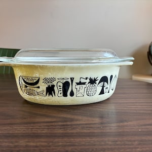 May include: A vintage yellow casserole dish with a black and white design of fruits, vegetables, and kitchen utensils. The dish has a glass lid.