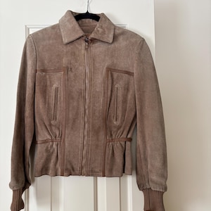 Puede incluir: Chaqueta de ante marrón con cuello y cremallera completa. La chaqueta tiene dos bolsillos verticales y puños acanalados. La cintura tiene un detalle fruncido. La chaqueta está colgada de una percha negra.