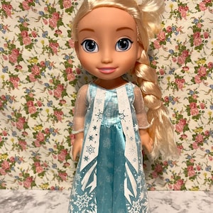 May include: A blonde doll wearing a blue dress with white snowflake designs. The doll has a long braid and is standing on a floral patterned background.