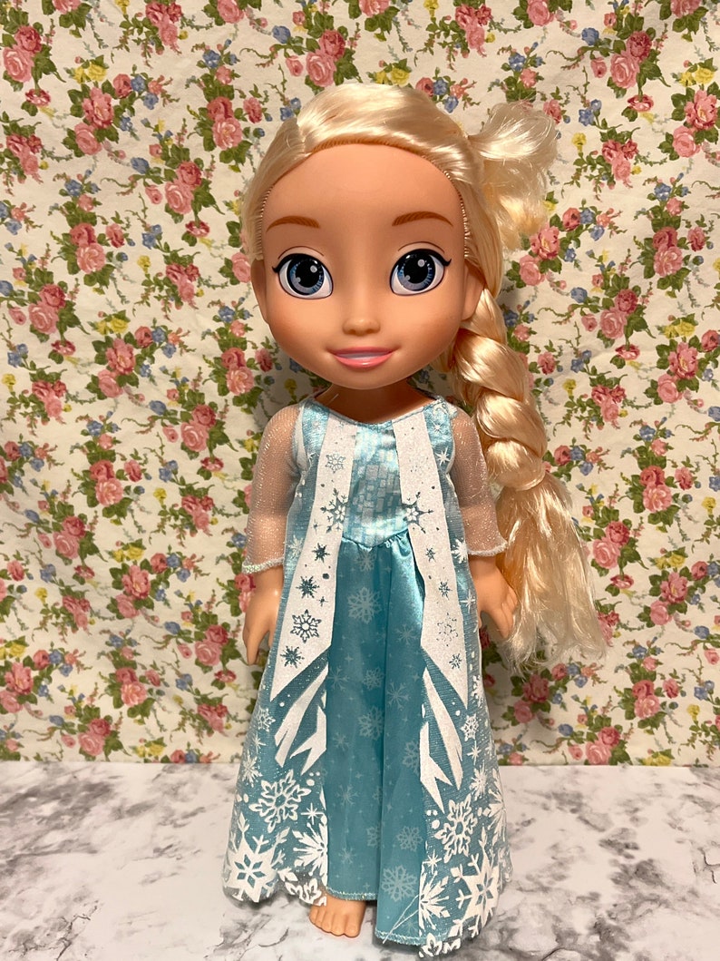 May include: A blonde doll wearing a blue dress with white snowflake designs. The doll has a long braid and is standing on a floral patterned background.