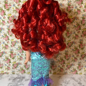 May include: A red-haired doll wearing a teal and purple mermaid costume. The doll has long, curly red hair and is wearing a teal dress with purple accents and a tulle skirt.