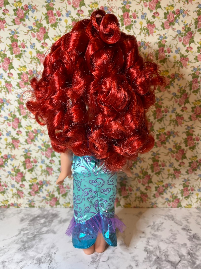 May include: A red-haired doll wearing a teal and purple mermaid costume. The doll has long, curly red hair and is wearing a teal dress with purple accents and a tulle skirt.