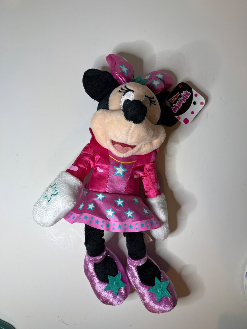 Pu&ograve; includere: Minnie Mouse di peluche rosa e turchese con un vestito rosa con stelle bianche e un fiocco turchese. Il peluche ha un'etichetta che dice "Disney Junior Minnie"
