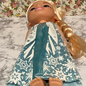 May include: A blue and white dress with snowflake patterns, worn by a doll with blonde hair and a smiling face.