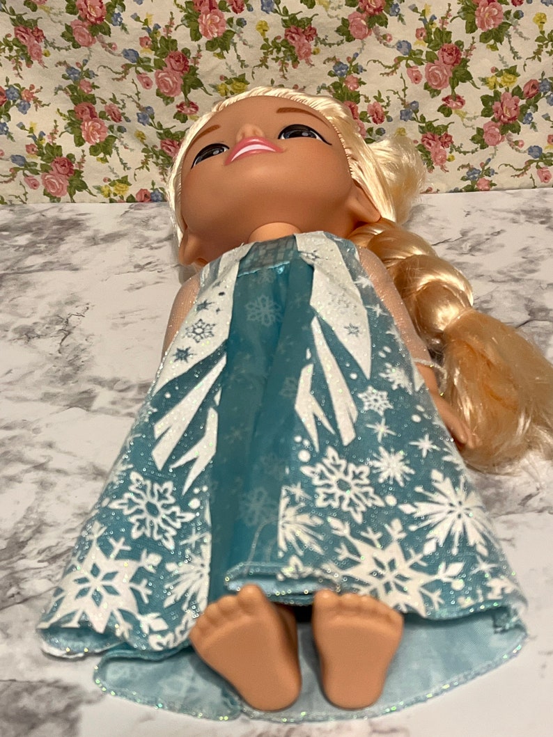 May include: A blue and white dress with snowflake patterns, worn by a doll with blonde hair and a smiling face.