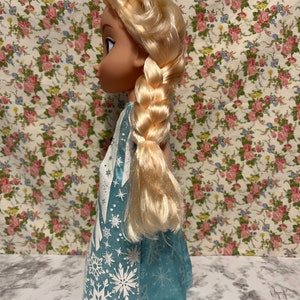 May include: A blue and white dress with a snowflake pattern. The doll has long blonde hair in a braid.