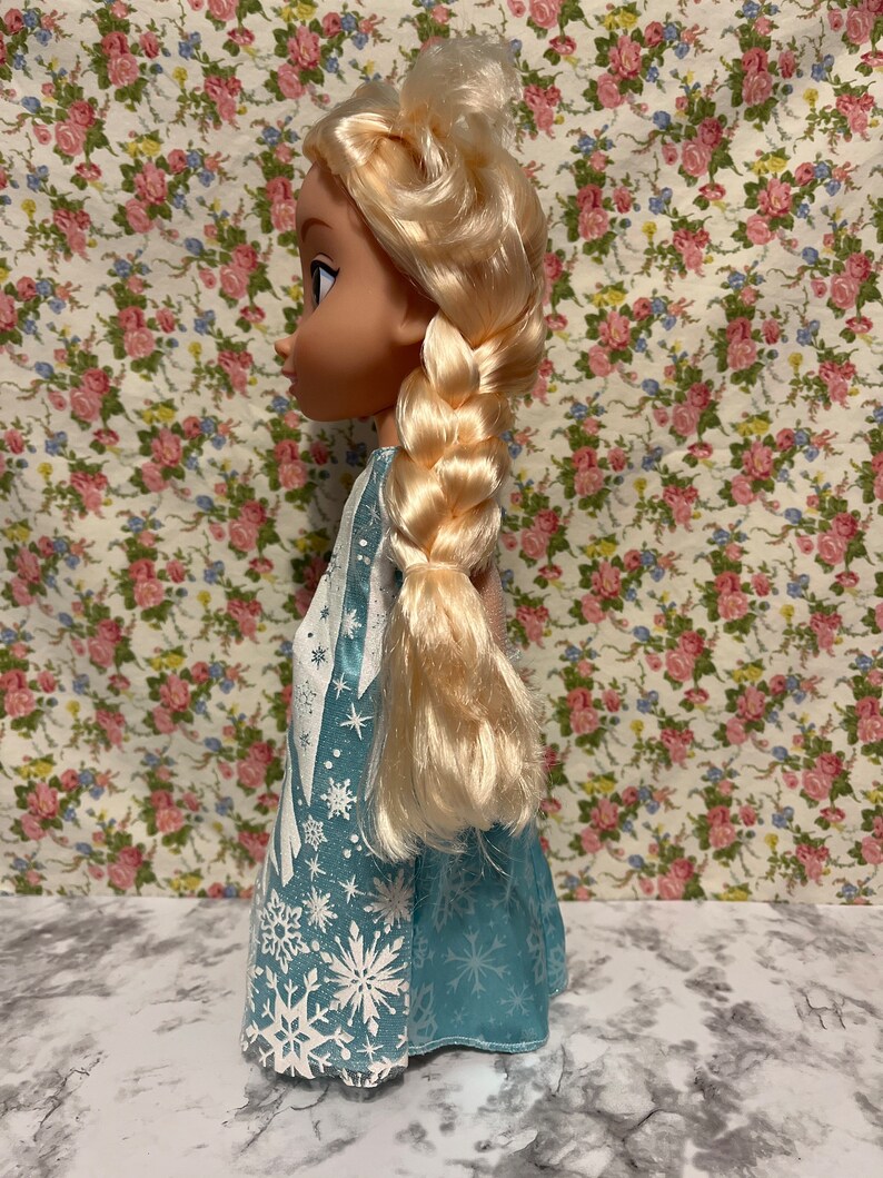 May include: A blue and white dress with a snowflake pattern. The doll has long blonde hair in a braid.