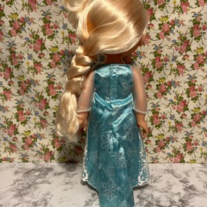 May include: A blue and white dress with a snowflake pattern. The dress has a long, flowing skirt and a fitted bodice. The doll has long blonde hair that is braided.