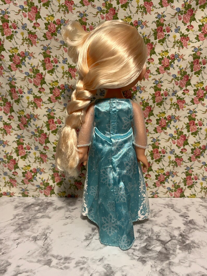 May include: A blue and white dress with a snowflake pattern. The dress has a long, flowing skirt and a fitted bodice. The doll has long blonde hair that is braided.