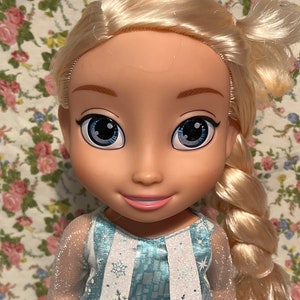May include: A blonde doll with blue eyes wearing a blue and white dress with snowflake designs. The doll has a long braid and is wearing a sheer blue top.