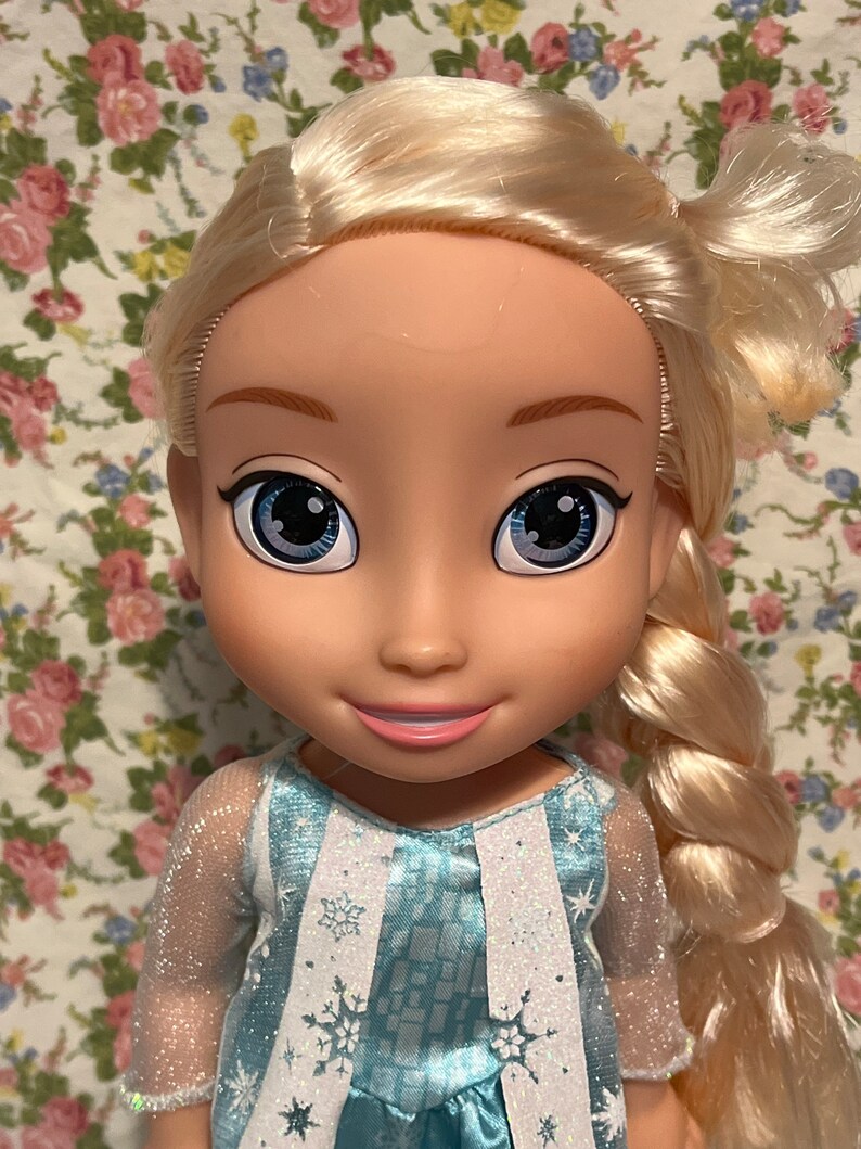 May include: A blonde doll with blue eyes wearing a blue and white dress with snowflake designs. The doll has a long braid and is wearing a sheer blue top.
