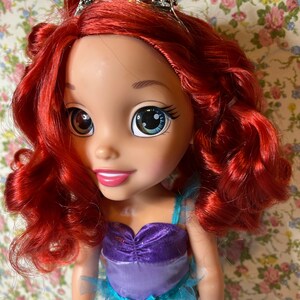 May include: A doll with red hair wearing a teal, purple and blue dress with a silver crown featuring a green seashell. The doll has large blue eyes and a friendly smile.