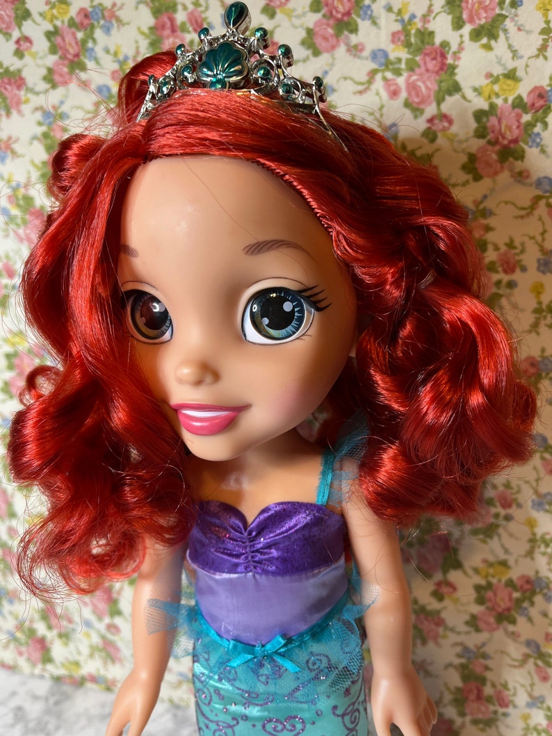 May include: A doll with red hair wearing a teal, purple and blue dress with a silver crown featuring a green seashell. The doll has large blue eyes and a friendly smile.