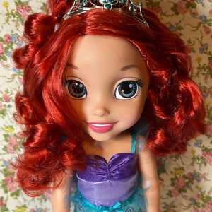May include: A doll with red hair wearing a teal and purple dress and a crown. The doll has large blue eyes and a bright smile.