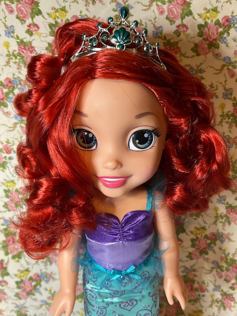 May include: A doll with red hair wearing a teal and purple dress and a crown. The doll has large blue eyes and a bright smile.