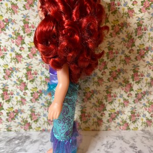 May include: A red-haired doll wearing a teal and purple mermaid costume with a crown on her head. The doll is standing on a white surface with a floral pattern background.