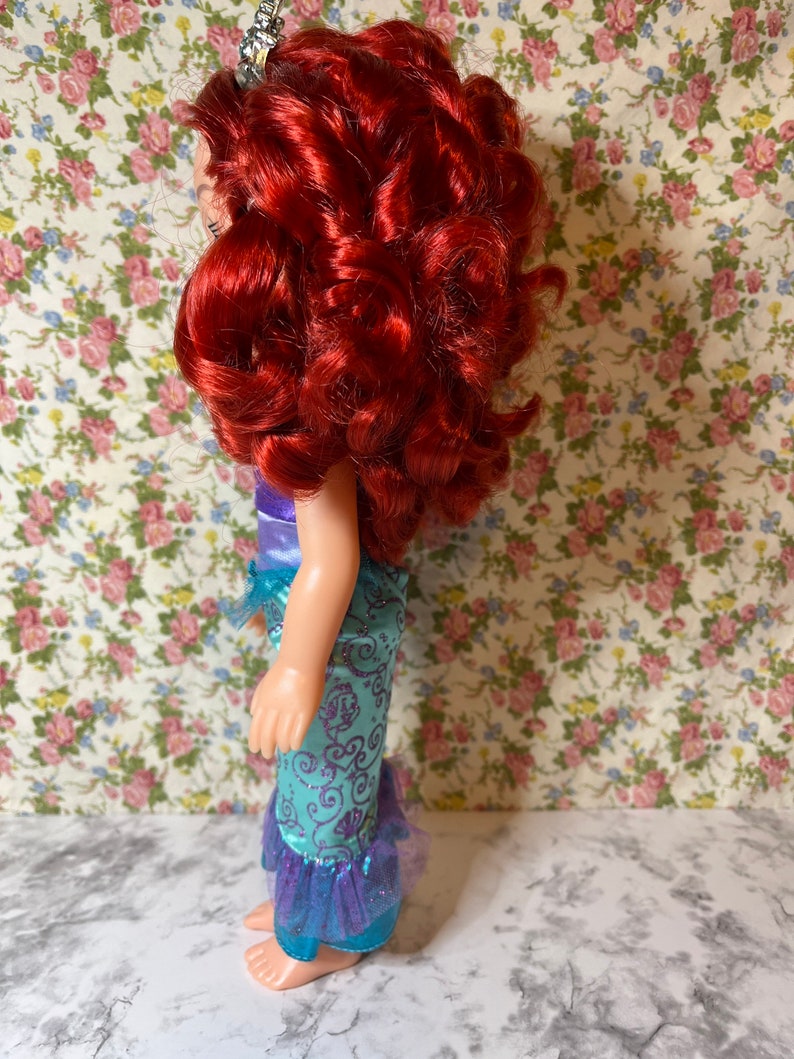 May include: A red-haired doll wearing a teal and purple mermaid costume with a crown on her head. The doll is standing on a white surface with a floral pattern background.