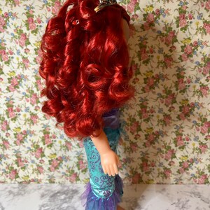 May include: A red-haired doll wearing a blue and purple mermaid costume with a sparkly crown. The doll is standing on a white surface with a floral pattern background.