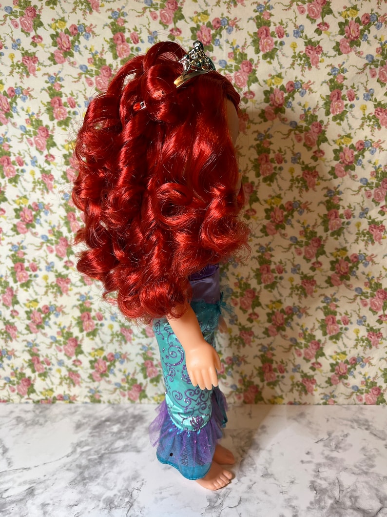 May include: A red-haired doll wearing a blue and purple mermaid costume with a sparkly crown. The doll is standing on a white surface with a floral pattern background.