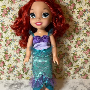 May include: A red-haired doll wearing a purple and teal mermaid costume with a crown. The doll has blue eyes and a smiling face.