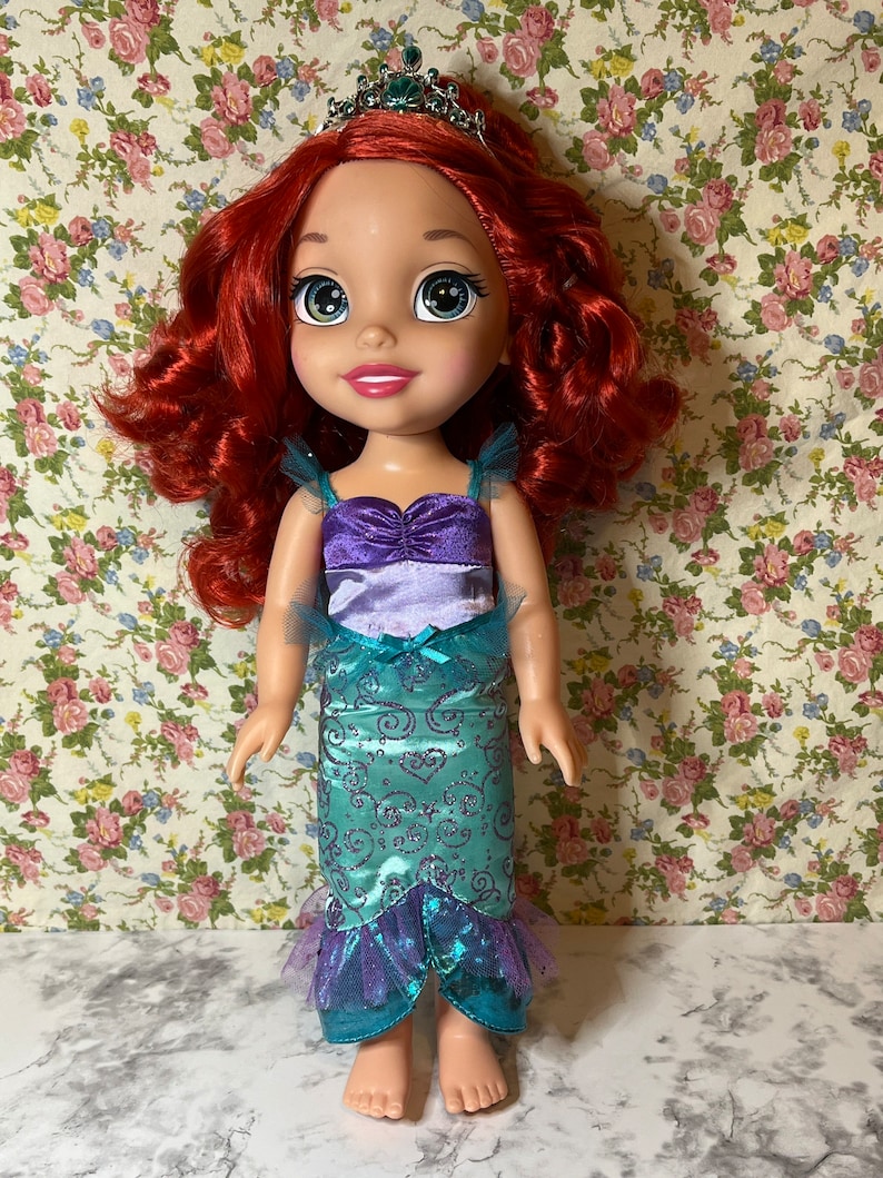 May include: A red-haired doll wearing a purple and teal mermaid costume with a crown. The doll has blue eyes and a smiling face.
