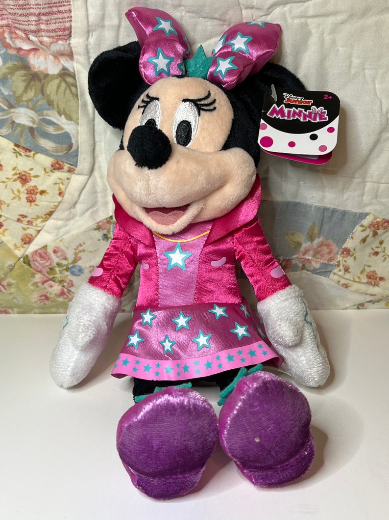 Pu&ograve; includere: Peluche rosa Minnie Mouse con un vestito rosa con stelle bianche e un fiocco rosa con stelle bianche. Il giocattolo ha un'etichetta che dice "Disney Junior Minnie" e "2+".