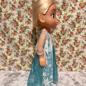 May include: A blue and white dress with a snowflake pattern. The doll has blonde hair and blue eyes.