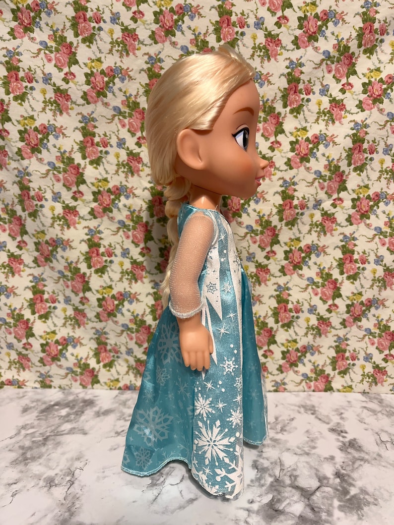 May include: A blue and white dress with a snowflake pattern. The doll has blonde hair and blue eyes.