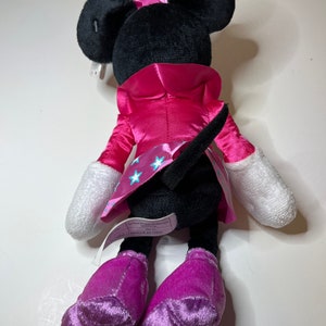 Pu&ograve; includere: Un giocattolo di peluche Minnie Mouse rosa e nero con un vestito di raso rosa con stelle bianche e scarpe viola. Il giocattolo ha una coda nera e un fiocco rosa.