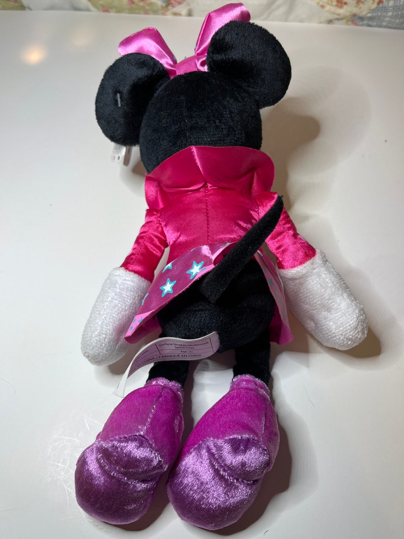 Pu&ograve; includere: Un giocattolo di peluche Minnie Mouse rosa e nero con un vestito di raso rosa con stelle bianche e scarpe viola. Il giocattolo ha una coda nera e un fiocco rosa.