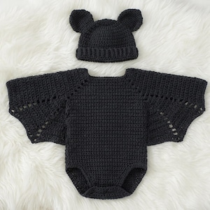 May include: A black crocheted bodysuit featuring wide, wing-like sleeves with openwork detailing and a matching black crocheted beanie hat with two rounded ears on top, both crafted with a consistent textured yarn stitch pattern.