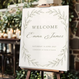 May include: A rectangular white wedding welcome sign on a wooden easel featuring a botanical leaf wreath border in olive green. The text reads WELCOME, Emma & James, SATURDAY 14 JUNE 2027, and CEREMONY AT FOUR O'CLOCK in a dark grey serif font.