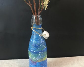 Puede incluir: Una botella de vidrio azul con un diseño de galaxia blanco y amarillo y salpicaduras de pintura blanca. La botella tiene un cierre de alambre metálico dorado con un tapón de cerámica blanco. La botella está llena de flores blancas.
