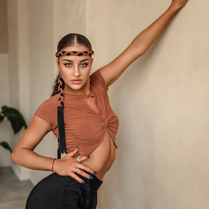 Puede incluir: Una mujer con un top marr&oacute;n, una diadema trenzada negra y marr&oacute;n, pantalones negros y esmalte de u&ntilde;as azul. Est&aacute; de pie con el brazo levantado contra una pared beige.