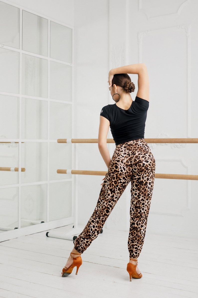 Puede incluir: Una mujer con una blusa negra y pantalones de estampado de leopardo posa en un estudio de baile. Est&aacute; de pie frente a una barra y un espejo. Lleva zapatos de tac&oacute;n marrones.