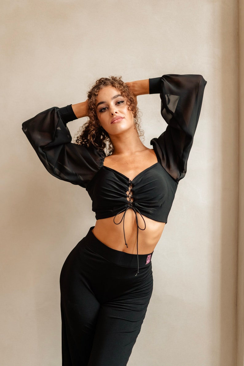 May include: A black crop top with long sheer sleeves and a lace-up detail at the front. The top is paired with black trousers.
