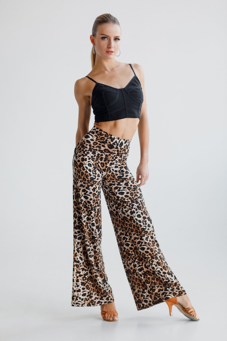 May include: A woman wearing a black crop top and leopard print trousers. She is standing with her legs slightly apart and her arms by her sides. She is wearing brown heeled sandals.