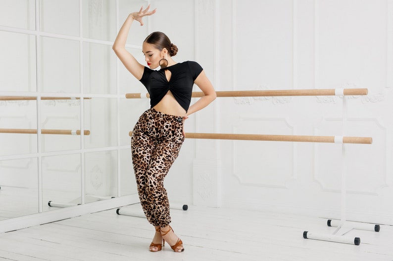 Puede incluir: Una mujer con un top negro corto con un detalle de torsi&oacute;n y pantalones de estampado de leopardo est&aacute; posando en un estudio de danza. Est&aacute; de pie frente a una barra de madera con soportes blancos.