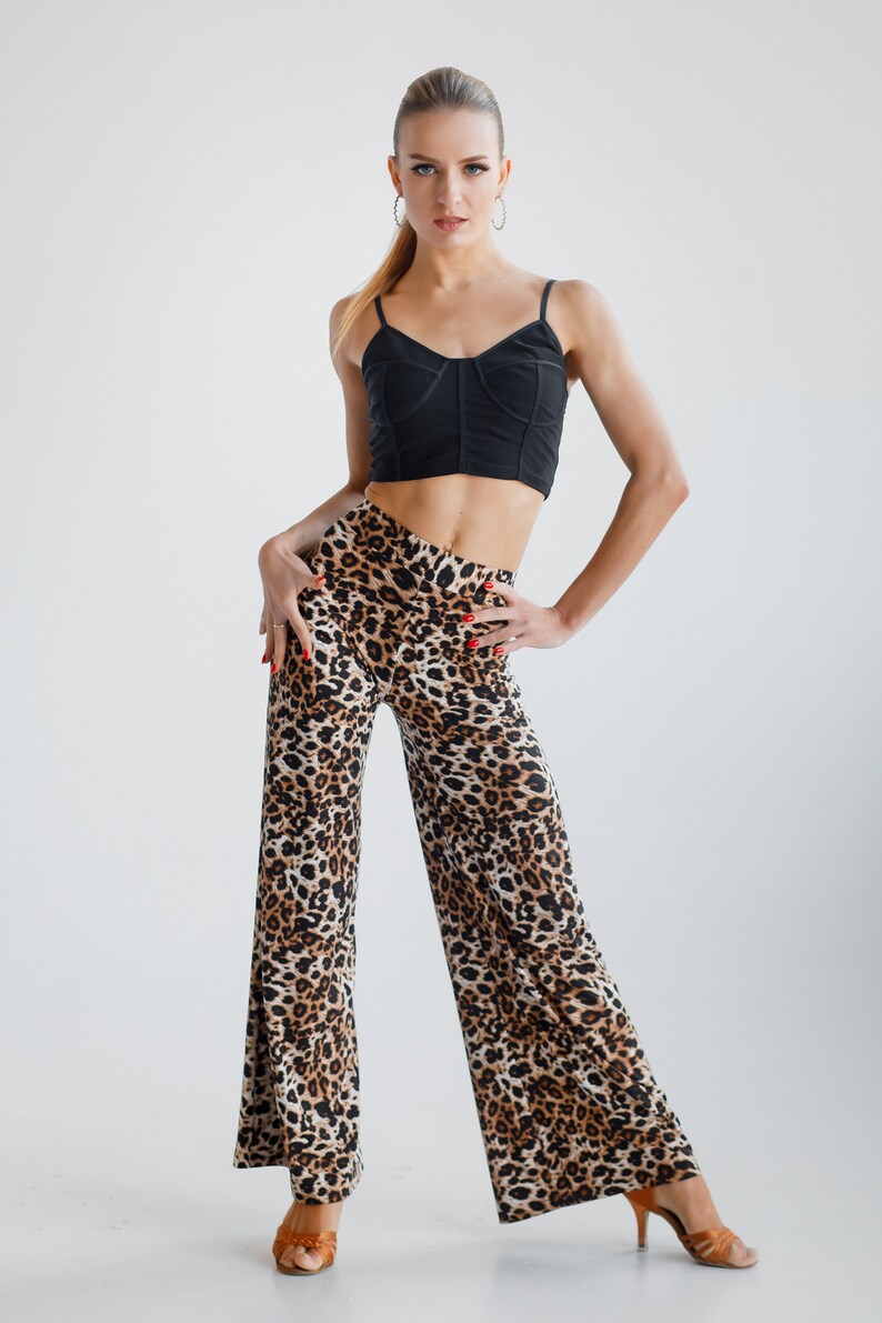 May include: A woman wearing a black crop top and leopard print trousers. She is standing with her hands on her hips and looking at the camera.