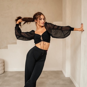 May include: A woman wearing a black crop top with a lace-up detail and black wide-leg trousers. She is standing in a room with white walls and a light brown floor.