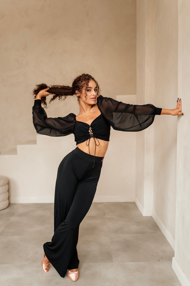 May include: A woman wearing a black crop top with a lace-up detail and black wide-leg trousers. She is standing in a room with white walls and a light brown floor.