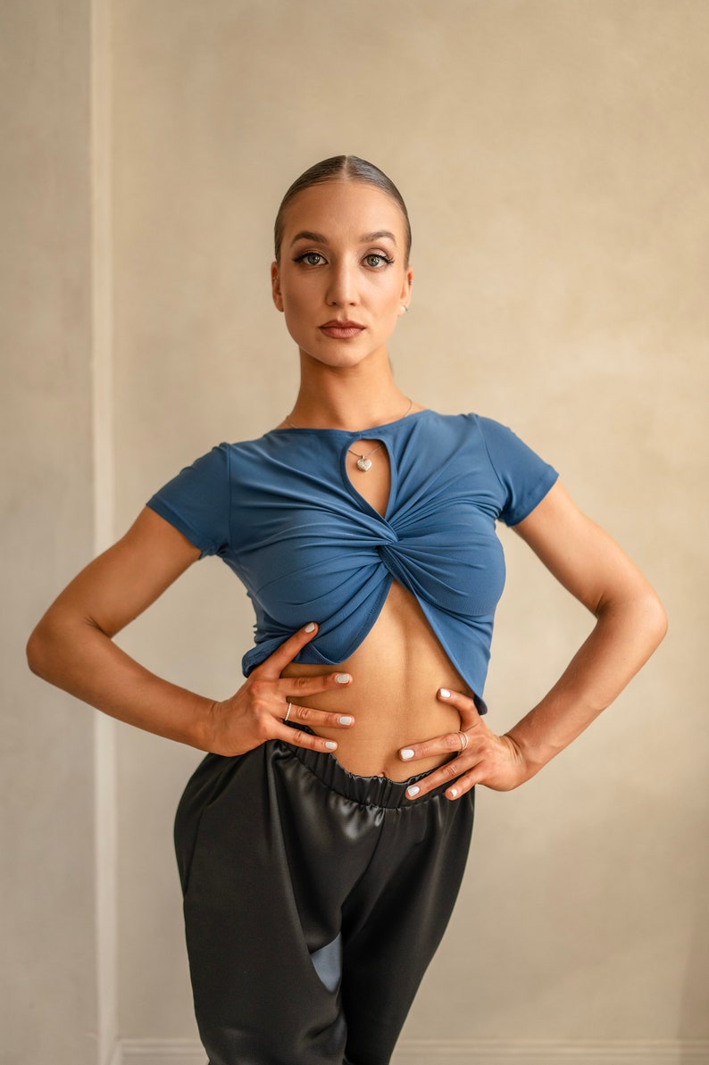 Puede incluir: Una mujer con un top azul corto con un detalle de torsi&oacute;n y pantalones de sat&eacute;n negro. Est&aacute; de pie con las manos en las caderas y mirando directamente a la c&aacute;mara.