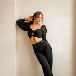 May include: A woman wearing a black crop top with a lace-up detail and black trousers. She is standing against a white wall and looking at the camera.