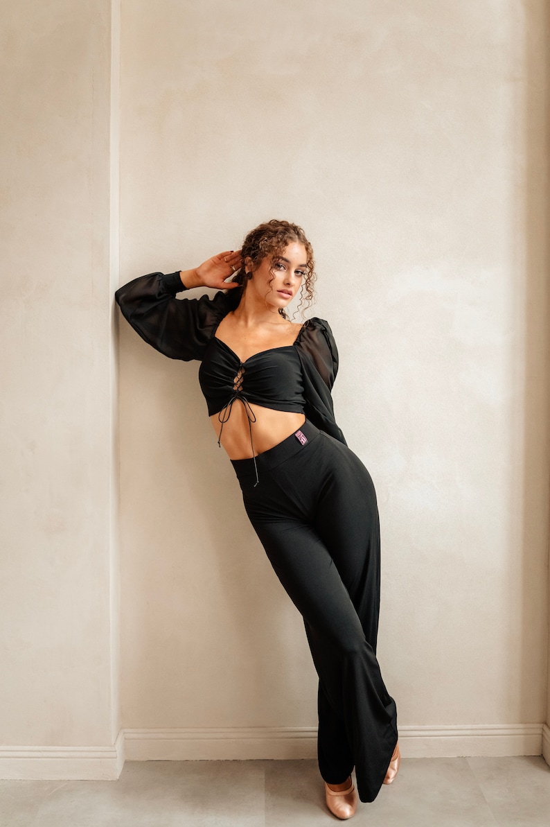 May include: A woman wearing a black crop top with a lace-up detail and black trousers. She is standing against a white wall and looking at the camera.