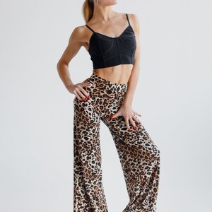 May include: A woman wearing a black crop top and leopard print trousers. She is standing with her hands on her hips and looking to the side. She is wearing brown heeled shoes.