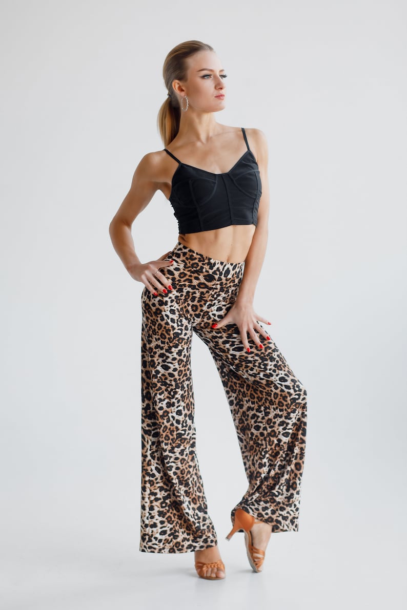 May include: A woman wearing a black crop top and leopard print trousers. She is standing with her hands on her hips and looking to the side. She is wearing brown heeled shoes.