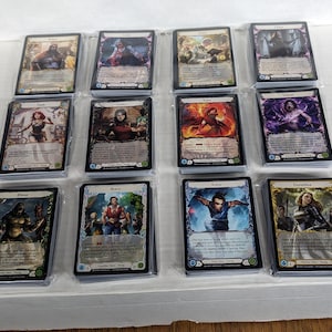 May include: A collection of trading cards, each in a protective sleeve. The cards feature fantasy art with characters in various poses and settings. The cards have a black border and are stacked in rows.