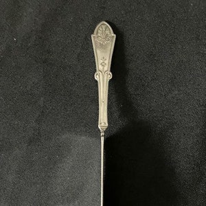 May include: A silver butter knife with an ornate handle. The handle features a decorative floral design at the top and a slender, curved blade. The knife is set against a dark background.