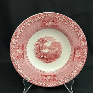 May include: A round, antique-style plate with a red and white design. The plate features a detailed landscape scene in the center, surrounded by a decorative border with floral and architectural elements. The plate is displayed on a clear stand against a black background.