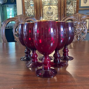May include: A set of six ruby red wine glasses with gold rims, displayed on a dark wooden table. The glasses feature a traditional goblet design with a rounded bowl and a delicate stem. The background shows antique wooden furniture.